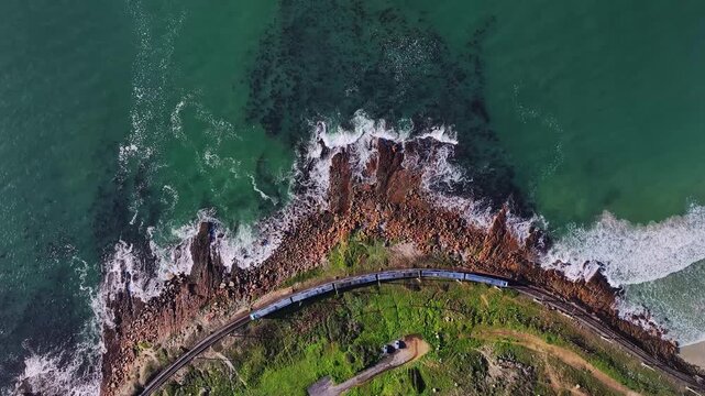 Aerial view of a train travelling along a coastal track with the ocean waves crashing against the rocky cliffs, Cape Town, Western Cape, South Africa.