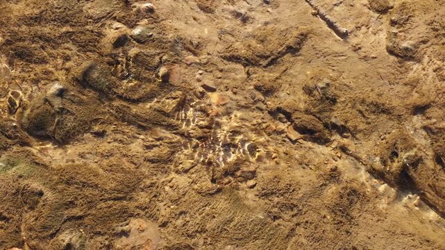 A detailed high-angle view captures clear, shallow water gently flowing over a rocky and sandy riverbed in Spain, with golden sunlight creating beautiful shimmering caustic patterns on the floor