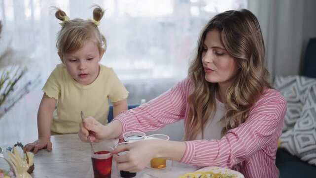 Mother and daughter create colorful art together. Child's hair is tied up in playful buns. Woman smiles warmly while guiding her. They sit at a bright kitchen table