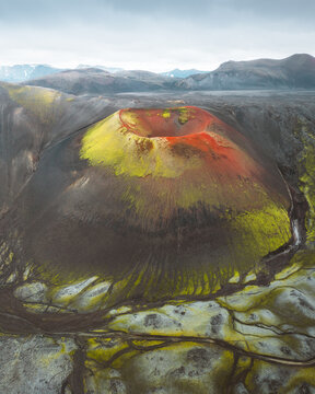 Aerial view of a volcanic crater, its dark slopes painted with streaks of yellow and red, contrasting with the grey landscape, Rangarthing ytra, Iceland.