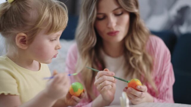 Mother and child paint colorful eggs together. Soft light shows calm, joyful moments. Child holds egg, concentrates. Mother guides, smiles gently. Perfect for Easter, family bonding, creativity themes