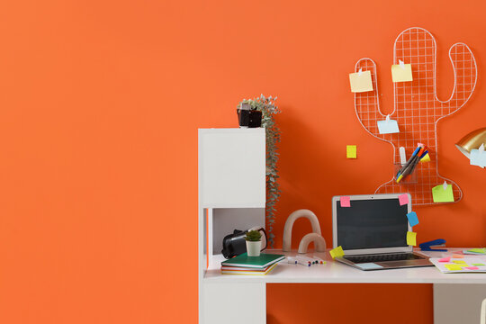 Workplace with blank laptop, sticky notes and pin board on orange wall in office