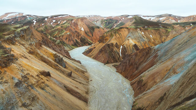 Aerial view of a muddy river snaking through a canyon of vibrant, ochre-streaked mountains, capped with patches of snow under a pale sky, Rangarthing ytra, Iceland.