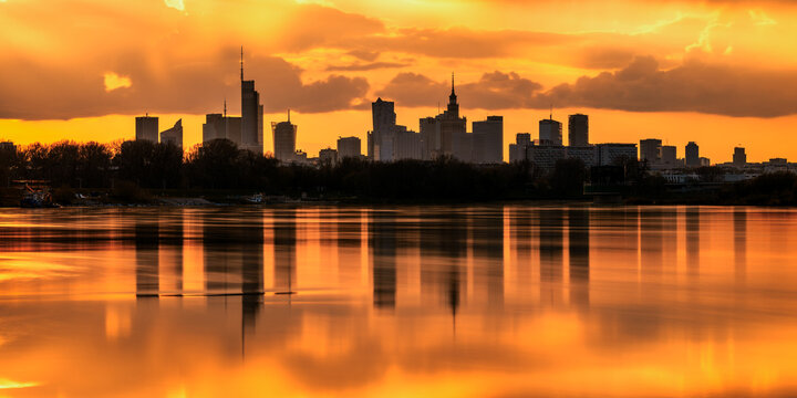 View of the city skyline mirrored in the tranquil water under a fiery sunset glow, silhouetting buildings against the bright sky, Warsaw, Masovian Voivodeship, Poland.