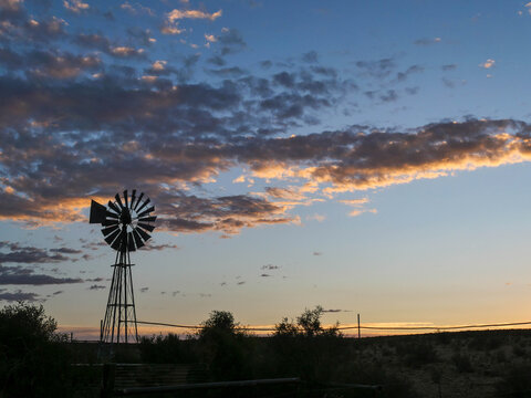 Windmill, blades spinning in the wind, pumping for water, in a Karoo field or rural farmland with clouds in the background,during sunset.