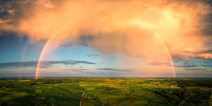 Aerial view of a vibrant double rainbow arcing across a lush green landscape under a dramatic, cloud-streaked sky, Stare Juchy, Warmian-Masurian Voivodeship, Poland.