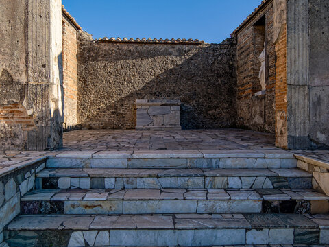 Ancient Roman sacred space in Pompeii featuring stone altar, marble columns, stone walls and terracotta roof fragments under blue sky. Marble steps lead to the preserved sanctuary near Naples.