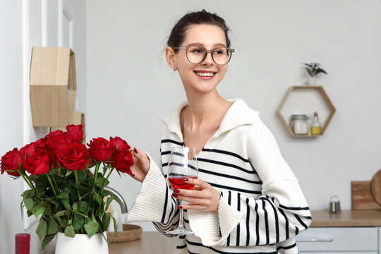 Beautiful young happy woman with glass of red wine and roses bouquet in kitchen