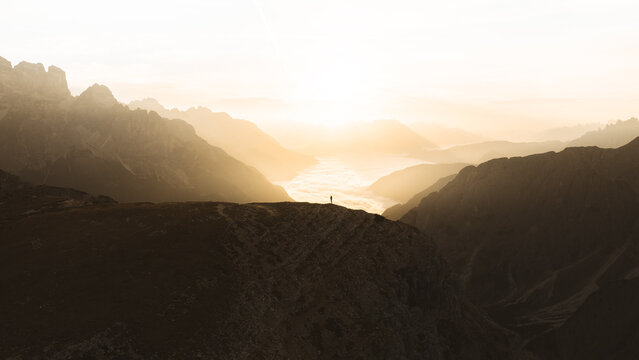 Aerial view of a solitary figure stands silhouetted against the golden light filtering through the rugged peaks of the Dolomites, Auronzo di Cadore, Veneto, Italy.