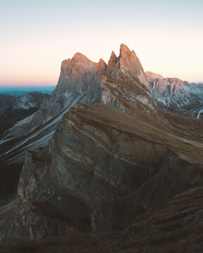 View of jagged peaks pierce the sky, bathed in the warm glow of sunrise over layered ridges, a symphony of earth tones and serene blues, Urtijei, Trentino-South Tyrol, Italy.