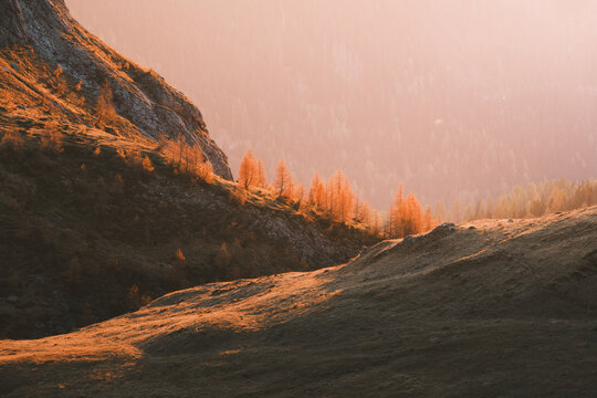 View of golden light kissing the rugged mountain slopes and vibrant autumnal trees casting shadows on the grassy terrain, San Vito di Cadore, Veneto, Italy.