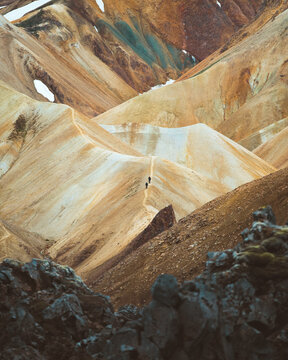 View of towering, multicolored mountains stripe the landscape with shades of ochre, cream, and teal, dwarfing two hikers in Rangarthing ytra, Iceland.