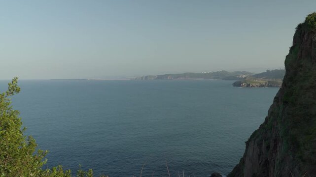 Stunning coastal cliffs rise above tranquil blue ocean waters near Candas, Asturias, Spain. Dramatic rocky coastline with distant islands under soft hazy sky. Beautiful Mediterranean seascape.