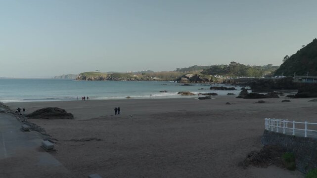 Scenic wide view of a sandy beach and rocky Atlantic coastline in Cand&aacute;s, Asturias, Spain. People walk along the shore while gentle waves reach the beach under clear skies.