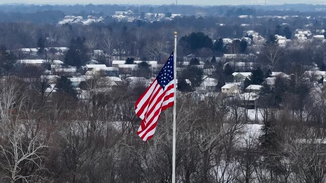 Patriotic American flag on flagpole slowly waving over America s suburb neighborhood in winter. Snowy houses and homes in background. Aerial view. Patriotic city of USA.