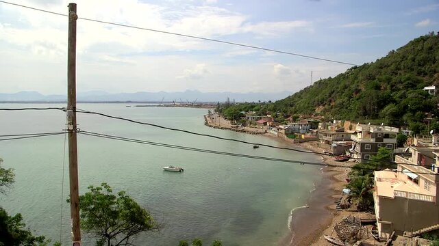Panoramic view of Cap-Haitien coastline in Haiti, with colorful hillside buildings along turquoise bay, small boat on water, distant mountains, power lines, and lush green hills under partly cloudy sk