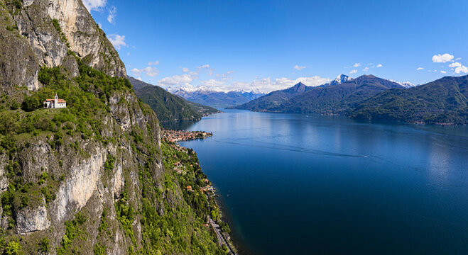 Aerial view of a small church perched on a rocky cliff overlooking the tranquil, dark waters of Lake Como, with snow-capped mountains in the distance, Domaso, Lombardy, Italy.