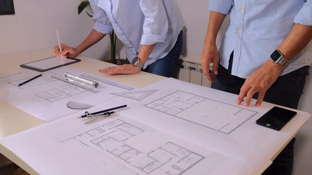 Two architects cooperating on building blueprint plans, the man measuring with a ruler and the woman working on a tablet with a stylus, in an architectural studio office