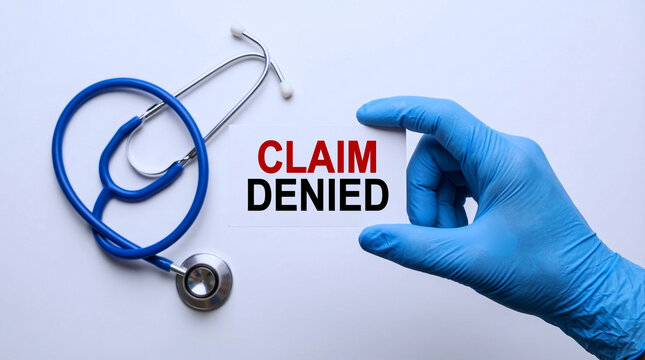 A gloved hand holds a card with the words claim denied next to a stethoscope on a white surface