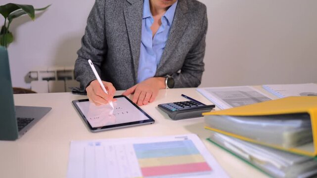 Professional accountant using a digital stylus pen to sign a document on a modern touchscreen tablet, reviewing financial data with a calculator and files on the office desk