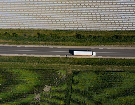 Aerial view of a lone white truck speeding along a dark asphalt road, juxtaposed against the geometric patterns of fields, Valensole, Provence-Alpes-Cote d'Azur, France.