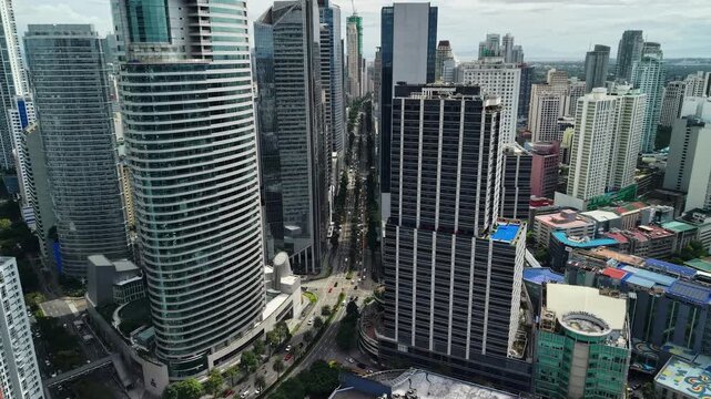 Drone view looks down a straight business district avenue framed by glass towers and heavy urban density as traffic moves below between modern high-rises in central Makati