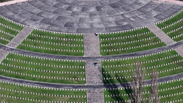 Aerial view of a circular war cemetery with rows of white crosses on green grass, gray stone paths and bare trees, Mittelwihr, Grand Est, France.