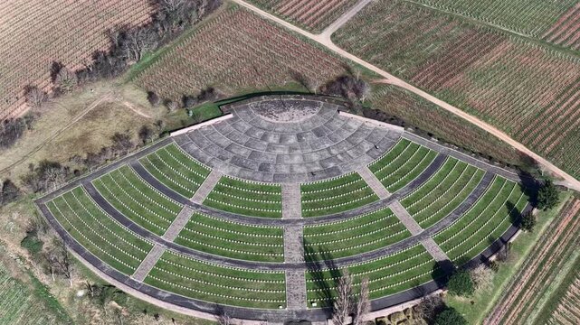 Aerial view of a fan-shaped cemetery with neat rows of white grave markers amidst green grass, contrasting with the surrounding brown vineyard, Mittelwihr, Grand Est, France.