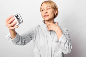 Mature woman with short blonde hair takes a selfie with a smartphone against a white background....