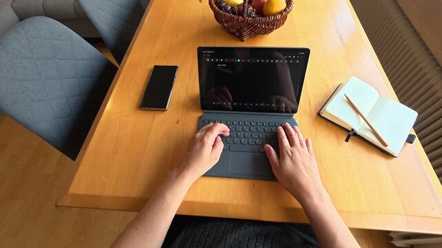 POV of a pregnant woman sitting at a dining room table at home typing on her tablet keyboard with her smartphone and notepad on the table.
