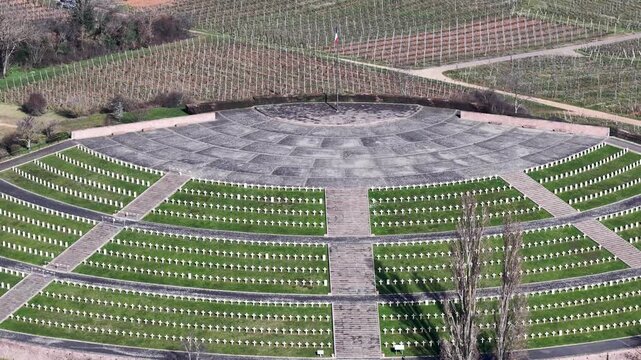 Aerial view of a solemn war memorial, with neat rows of white crosses against green grass, surrounded by vineyards, Mittelwihr, Grand Est, France.