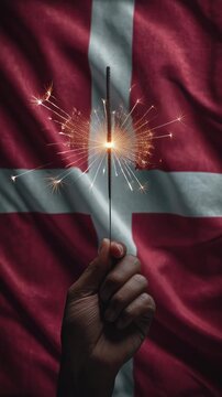 Hand holding a lit sparkler with sparks flying against a red and white flag background.