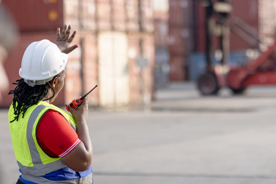 African woman engineer with safety vest and helmet holds worky talky, gives hand signals to stop forklift at container yard. Female foreman worker working at logistic shipping cargo container yard.
