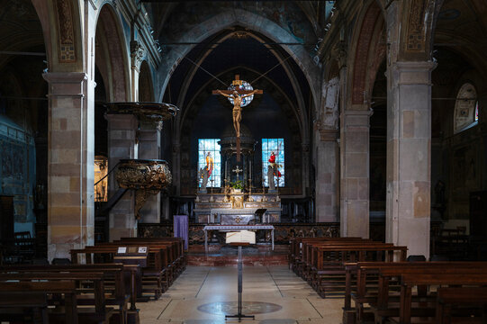 View inside a historic Catholic church in Angera, Italy, featuring wooden pews, a central aisle, altar and large crucifix illuminated by stained glass windows.