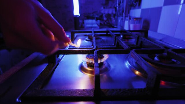 Male hand turns on gas stove, igniting two burners with a match in a dark kitchen. Close-up shot in dramatic blue light with a slow-moving camera.