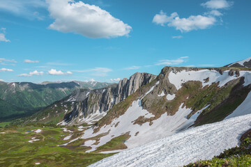 Scenic view from grassy flowering cliff along ridge with snow cornice to sharp rocks and rocky sheer crags under lush clouds in blue sky. Sunlight and shadows in high mountains in changeable weather. © Daniil