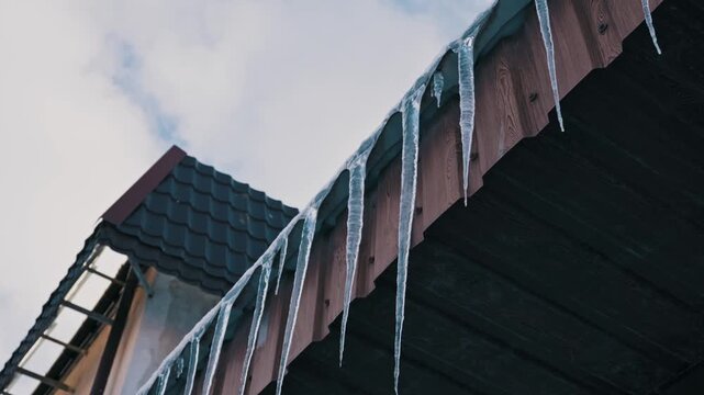 Long sharp icicles hang in a row from the wooden eaves of a house roof. A low-angle timelapse captures clouds moving quickly across the cold blue sky.