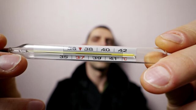 Man hands hold a glass mercury thermometer showing a high body temperature of 40.5 degrees Celsius. The sick man in the background is out of focus, creating a point-of-view perspective.
