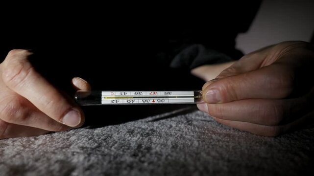 Male hands hold a glass mercury thermometer showing a high fever reading. The scene is dark with dramatic lighting, creating a somber mood of sickness.