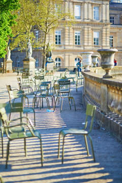 Scattered empty green chairs casting dramatic shadows on a sunlit gravel path in a historic Parisian garden, with classical statues and old architecture in the background