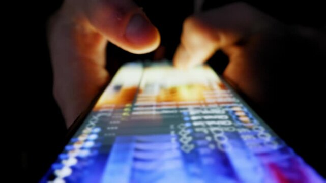 Hands scroll through an endless social media feed on a smartphone. The screen illuminates the fingers in an otherwise completely dark room.