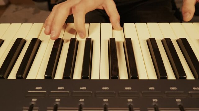 Musician hands play a melody on a MIDI synthesizer keyboard in a recording studio. Close-up shot focuses on the finger movements across the black and white keys with warm lighting.