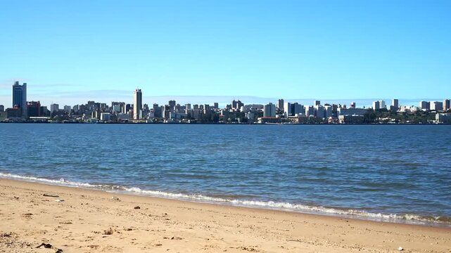 Panoramic skyline of Maputo, Mozambique across Maputo Bay from beach, modern high-rises and mid-century buildings on clear blue day, sandy shore foreground.