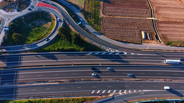 Aerial view of highway lanes merging near fields. Drone shot showing traffic on a multi lane road next to cultivated vegetable gardens.