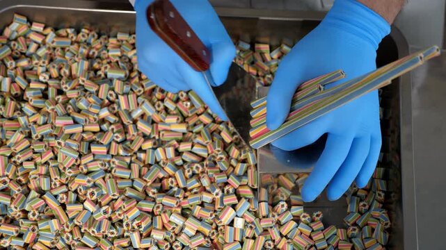 Close-up of a professional pastry chef slicing long multicolored caramel candies with a pattern inside. He uses a metal spatula in the production process of making sugar candies at a confectionery.