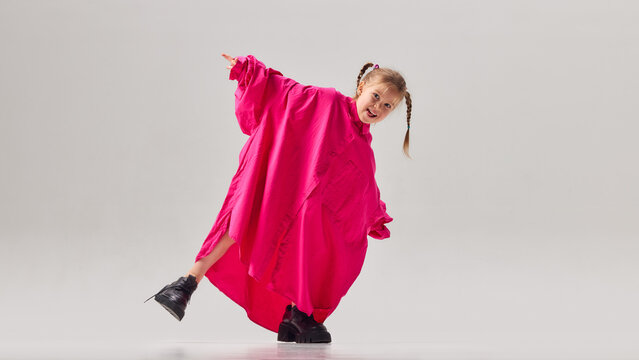 Little girl in oversized pink shirt and large shoes posing playfully in studio. Concept of childhood imagination, playful fashion styling, growing up and kids exploring adult roles.