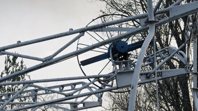 Blades of a deactivated carousel motor spin slowly from the wind in a deserted park. Overcast sky and bare trees create a melancholic and post-apocalyptic atmosphere.