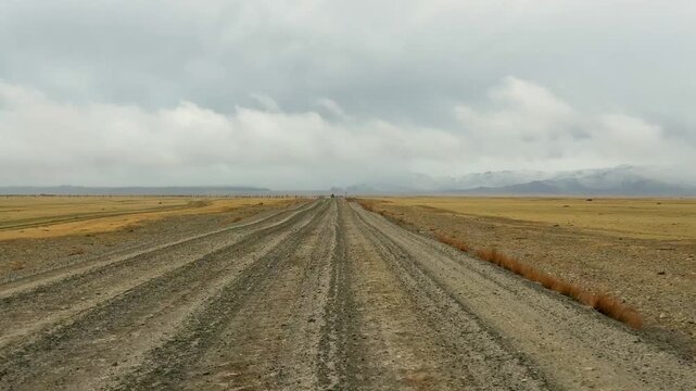 Gravel road stretching from Altai towards the Mongolian border with snowy mountain ridge. Desert steppe landscape under stormy cloudy sky showcasing wild remote terrain