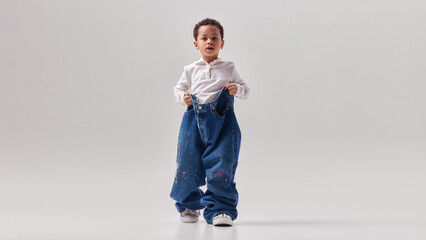 Little boy wearing oversized denim trousers and casual shirt standing in studio. Concept of childhood imagination, playful fashion styling, lifestyle creativity and growing up ideas. © Lustre Art Group 
