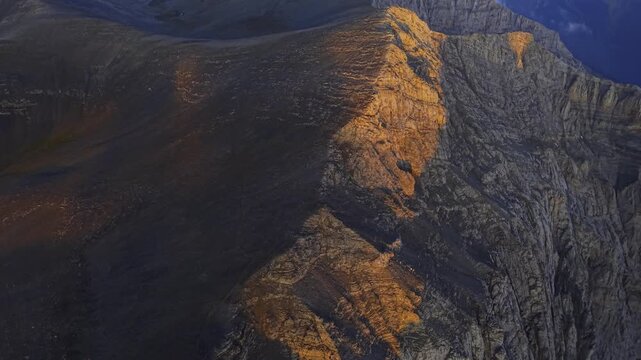 Aerial view of rocky peaks, where sunlight bathes the rugged terrain in a warm, golden glow, creating a striking contrast with the shadowy depths, Mount Olympus, Larissa, Greece.